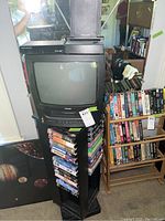Toshiba 13-inch CRT TV with DVD player on top of a black rotating VHS tape rack filled with multiple VHS tapes. Adjacent wooden rack also full of VHS tapes.