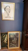 Three framed pieces of wall art placed against a dark blue wall and wooden cabinet. One is hanging and two are resting below it. All depict abstract human faces or figures in a monochromatic palette.