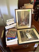 Photo showing two framed wall art pieces leaning on a drawer, one with a city street scene and another with architectural features, surrounded by other art prints and papers