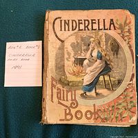Front cover of Cinderella Fairy Book from 1891, showing a colorful illustration of Cinderella sitting by a fireplace, with some wear on the cover.
