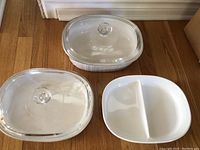 Three white ceramic Corning Ware casserole dishes with classic ridged exterior design, two with clear glass lids, one divided with two sections.