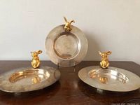 Three silver-plated round dishes with bronze rabbits attached on rim, shown from front angle on wooden table.
