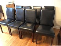 Front view of eight black bicast leather dining chairs arranged in two rows on a hardwood floor.