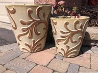 Two beige clay plant pots with raised brown leaf pattern placed outdoors on stone pavers.