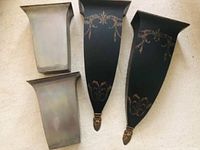 Pair of black tapered metal wall sconces with gold scroll design and removable interior containers on floor.