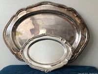 Photo showing both silver-plated trays stacked, larger tray in back with scalloped edge and smaller oval tray in front with decorative edge