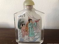 Front view showing one painted scene of two women inside the clear glass bottle with a cork stopper on top.