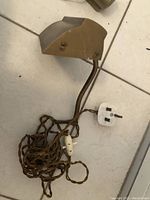 Three picture light fixtures laid on floor showing their form, plugs, and wiring. Two longer cylindrical lights with antique gold finish, mounting wires, and one smaller angled brass-finished light with brown twisted fabric cord and a British plug.