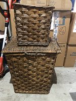 Large rectangular wicker laundry hamper with hinged lid stacked with smaller square-shaped wicker waste paper bin on top. Both items are brown with a woven pattern, showing wear.
