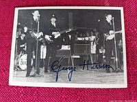 Black and white photo card showing The Beatles performing live, with George Harrison's printed signature at the bottom center.