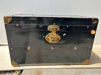 Front view of black metal steamer trunk showing brass latch and corner protectors, minor surface scratches visible.