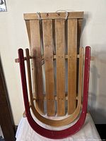 Front angled view of vintage child's wooden sled showing red curved runners, natural wood slatted seat, and white pull rope attached to front crossbar.