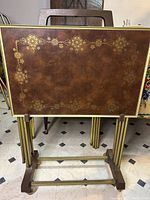 Front view of one brown vintage TV tray table panel showing detailed gold floral pattern, gold metal legs, and part of wooden stand beneath.