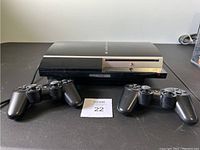 Front view of PS3 console with 2 controllers placed side by side in front on black surface.