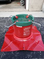Close-up photo of the metal Christmas tree stand showing green top with four red knobs and tightening screws, red weighted base with angular shape for stability.