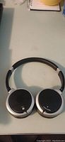 Over-ear headphones shown flat on surface, black and silver with cushioned ear cups and adjustable headband.