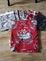 Photo showing folded cotton and linen fabric pieces with various patterns laid on wooden floor, including red toile pattern with figures, black paisley, and red and cream paisley.