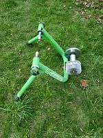 Green metal frame bicycle trainer with resistance unit placed on grass, showing side view and structure.