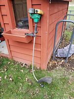 Green and yellow gas-powered edge trimmer leaning against wood wall outdoors.
