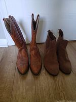 Pair of boots showing brown leather cowboy boots on left and brown suede heeled boots on right on wood floor.