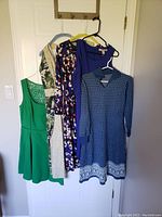 Five women's dresses hanging on hangers on a door, showing full length and variety of colors and styles