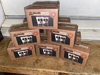 Twelve brown cardboard boxes containing sets of two shot glasses each, stacked and displayed on wooden surface with branding and product image visible