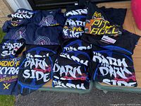 Photo showing multiple folded black T-shirts with The Dark Side Star Wars themed graphic text and symbols, neatly folded on the ground.