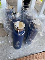 Six new metal beverage coolers with blue matte finish and silver lids, all wrapped in plastic packaging, arranged together on concrete floor.