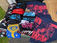 Overview photo showing 26 folded new men's medium T-shirts with various music band logos and themes, mostly black with colorful prints.