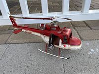 Photo of metal helicopter model with red and white coloring, showing side view on pavement with white fencing background.