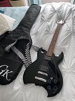 Full view of black Lyon by Washburn electric guitar lying on white bedspread next to soft carry case with checkered strap and logo