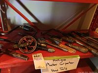 Various antique carpenter hand tools laid out on a shelf including hand drills, chisels, spokeshave, and marking gauge.
