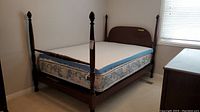 View of vintage wood four post bed with mattress and mattress topper against white walls and carpet floor.