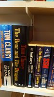Shelf with hardcover and paperback fiction books, featuring multiple Tom Clancy titles including 'Support and Defend' and 'The Bear and The Dragon'.