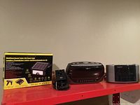 Photo showing multifunctional solar LED fixed security light box, HP webcam with attached USB cable, Nextech alarm clock radio, and Sanyo alarm clock radio from left to right.