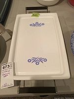 White rectangular ceramic broil bake tray with blue cornflower floral designs on top, set in a metal stand with black handles.