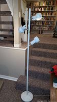 Full view of the floor lamp showing the three adjustable white spotlights on a white metal pole with the base on stairs.