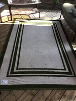 Full view of beige textured outdoor rug with green triple stripe border, laid on wooden deck with some natural debris on surface.