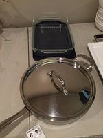 Photo showing the Pyrex glass lasagna dish with black plastic cover and the large stainless steel Wolfgang Puck skillet with lid on a table.