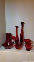 Photo showing 9 vintage red and cranberry glass pieces on display including vases, pitchers, bowls, and a stemmed glass.