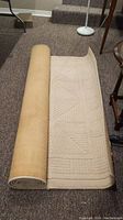 Partially unrolled beige textured rug on carpeted floor showing back side and edge detail.