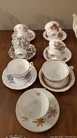 Photo showing 7 vintage floral teacup and saucer sets with various floral patterns and gold trim on a wooden table.