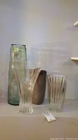 Four vases displayed side by side on a shelf including a tall green cylindrical glass vase, a clear fluted glass vase, a greyish-brown ceramic vase, and a clear flared glass vase.