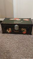 Front view of vintage metal trunk showing dark color, affixed old travel stickers, and metal latch lock.