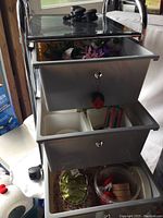 Full view of the three-bin plastic storage trolley with items inside bins visible, metal frame with wheels, and chrome knobs on bins.