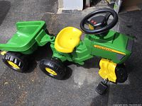 Photo of the full John Deere pedal tractor with attached green wagon, showing side view including seat, steering wheel, and large rugged wheels.