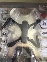 Drone in package showing front view of black folding drone with four propellers and camera.