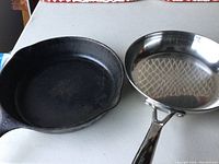 Two frying pans placed side by side on a table: a black cast iron pan showing wear and a shiny stainless steel pan.
