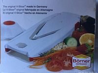 Original Börner V-Slicer showing the white base with V-shaped stainless steel blade, surrounded by sliced vegetables and fruits, with clear branding and 'made in Germany' text.