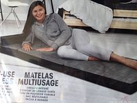 Photo showing a woman laying and sitting on the black rectangular foam mattress inside a room with wooden floors and modern furniture.